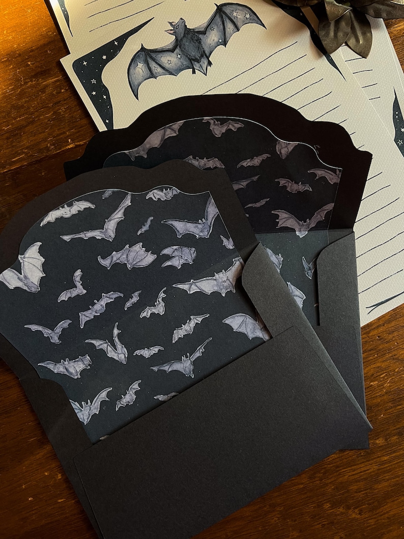 May include: Black stationery set featuring bat illustrations. The set includes envelopes with a bat pattern, lined paper, and a decorative bat illustration. The envelopes are black, and the paper has a starry border.