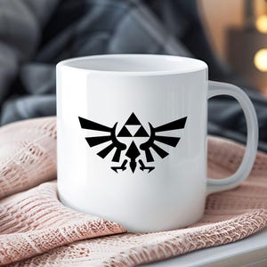 May include: White ceramic mug with a black emblem of a triangle above a stylized bird with wings. The mug has a curved handle and is set against a soft, textured background. The background is a soft pink.