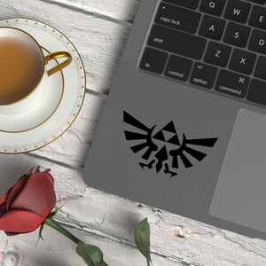 May include: A black Legend of Zelda crest sticker is on a laptop. A cup of tea in a white teacup with gold detailing sits on a matching saucer. A red rose rests on a white wooden surface.
