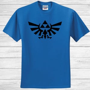 May include: A royal blue t-shirt with a black emblem design. The emblem is a stylized symbol with a central triangle and wing-like shapes. The t-shirt has a crew neck and short sleeves.
