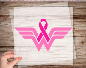 May include: A pink and white Wonder Woman logo with a pink ribbon in the center. The logo is on a clear sheet, ready to be pressed onto a surface.