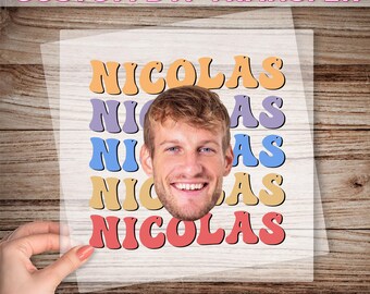 May include: A custom DTF transfer featuring the name "NICOLAS" in various colors, with a smiling face in the center. The text "READY TO PRESS" is at the bottom. The transfer is held by a hand.