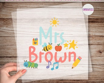 May include: A clear DTF transfer with the text "Mrs. Brown" in a playful font, accompanied by colorful illustrations of a sun, bee, apple, stars, and pencil. The words "CUSTOM DTF TRANSFER" and "READY TO PRESS" are also visible.