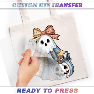 A hand is holding a piece of paper over a white tote bag with a Halloween-themed design featuring a ghost, pumpkin, and bow.