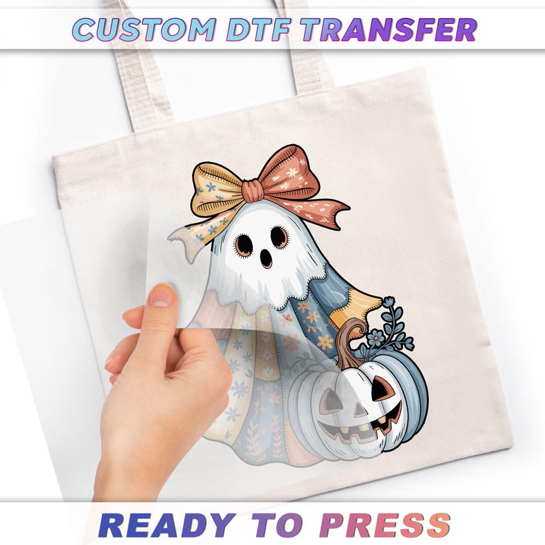 A hand is holding a piece of paper over a white tote bag with a Halloween-themed design featuring a ghost, pumpkin, and bow.