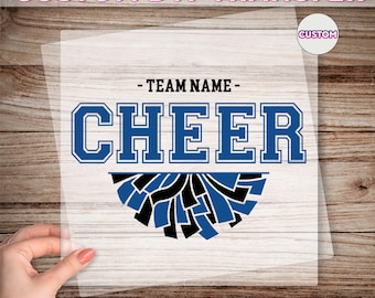 May include: A clear DTF transfer sheet with the word "CHEER" in blue block letters. The text "- TEAM NAME -" is above the word "CHEER". Below the word "CHEER" is a pom-pom design in blue and black. The text "CUSTOM DTF TRANSFER" and "READY TO PRESS" are also visible.