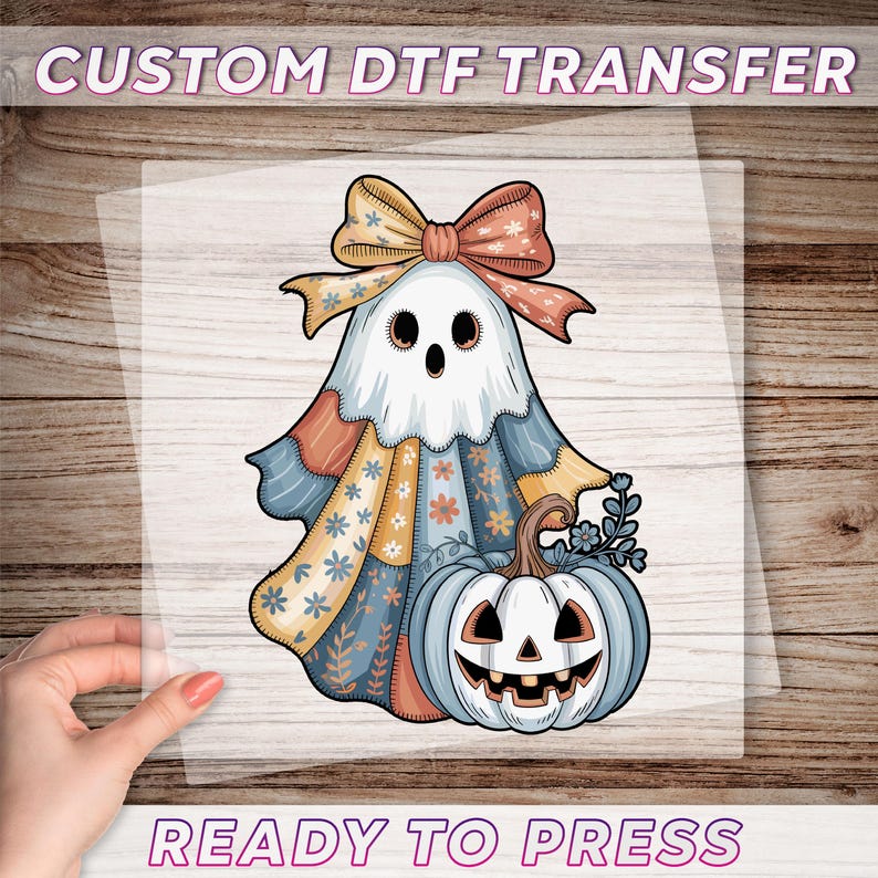 a hand holding a piece of paper with a Halloween-themed illustration of a ghost, pumpkin, and bow on it.