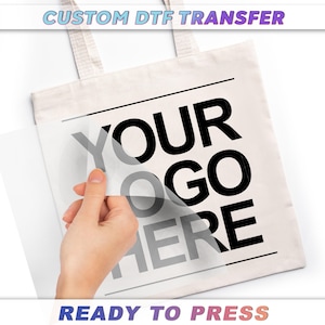 A hand is holding a white tote bag with the text "YOUR" and "GO HERE" printed on it, and the text "CUSTOM DTF TRANSFER" is visible above the bag.