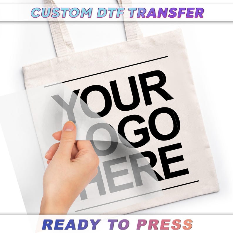 A hand is holding a white tote bag with the text "YOUR" and "GO HERE" printed on it, and the text "CUSTOM DTF TRANSFER" is visible above the bag.