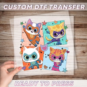 May include: A clear sheet featuring a colorful design of four cartoon superhero cats. Each cat is in a different pose and costume, with stars and hearts around them. The text "CUSTOM DTF TRANSFER" and "READY TO PRESS" are at the top and bottom.