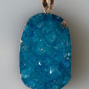 Peut inclure: Pendentif rectangulaire avec une bélière et une bordure dorées. Le pendentif présente une pierre bleu-vert cristalline et vibrante. La pierre a une surface rugueuse et texturée, sertie dans un métal doré.