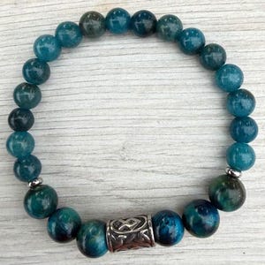 May include: A beaded bracelet featuring round, polished blue-green apatite gemstones arranged in a circular pattern. The bracelet includes a single cylindrical silver-toned metal bead with an intricate embossed pattern and two small silver-toned spacers.