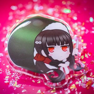 Puede incluir: Un amuleto de acrílico transparente con un personaje de anime de dibujos animados con cabello negro, ojos rojos y un cuello blanco. El personaje sostiene un cuchillo y está colocado sobre un fondo rosa con purpurina iridiscente.