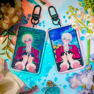 May include: Two clear acrylic keychains featuring a colorful anime-style illustration of a character in a red jacket, white shirt, and holding hands. Each keychain has a black clasp and a phone-shaped design. The background is a mix of blue and green.