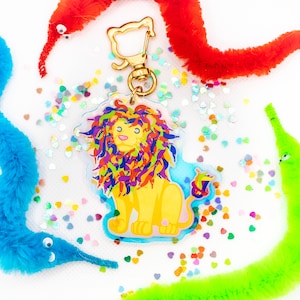 May include: A colorful rainbow lion keychain with a gold chain and a sparkly background. The lion is surrounded by colorful, fuzzy, worm-like toys.