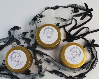 May include: Three gold massage candles with white labels reading "Sexy", "Tastee", and "Lace". The labels also feature the text "Praise Worthy Candles". A black lace garment with bows is draped around the candles.