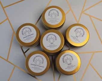 May include: A top-down view of seven gold candle tins with white labels. Each label reads "Praise Worthy Candles" and lists a scent, such as "Sexy" and "Tastee". The background is a white surface with a gold geometric pattern. Each tin is approximately 6 cm in diameter.