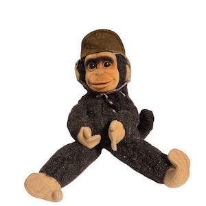 May include: A vintage plush monkey doll with a brown cap and ear coverings. The monkey has a tan face, hands, and feet, and is wearing a dark brown outfit. The doll is sitting with its legs spread.