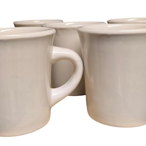 May include: A collection of five off-white ceramic mugs. Each mug has a curved handle and a slightly flared rim. The mugs are arranged in a group, with some overlapping, against a white background.