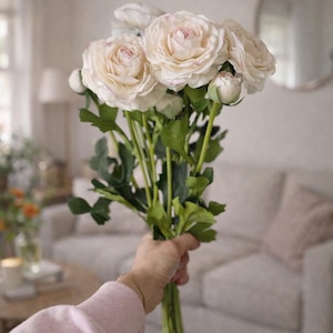 May include: A bunch of pale pink and white ranunculus flowers with green stems and leaves. The flowers are held in a hand, with a blurred background of a living room.