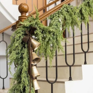 Puede incluir: Una guirnalda festiva para escaleras con ramas de hoja perenne artificiales, campanas doradas y una cinta de terciopelo marrón. La guirnalda está colocada sobre una barandilla de madera y balaustres de metal negro.
