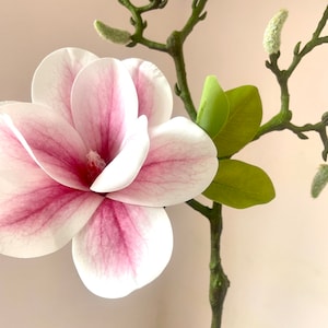 Puede incluir: Un solo tallo de flores de magnolia artificiales con p&eacute;talos rosados y blancos y hojas verdes.