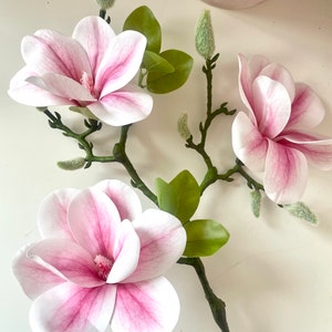 Puede incluir: Una rama de flores de magnolia artificiales con tres flores rosas y blancas y hojas verdes.
