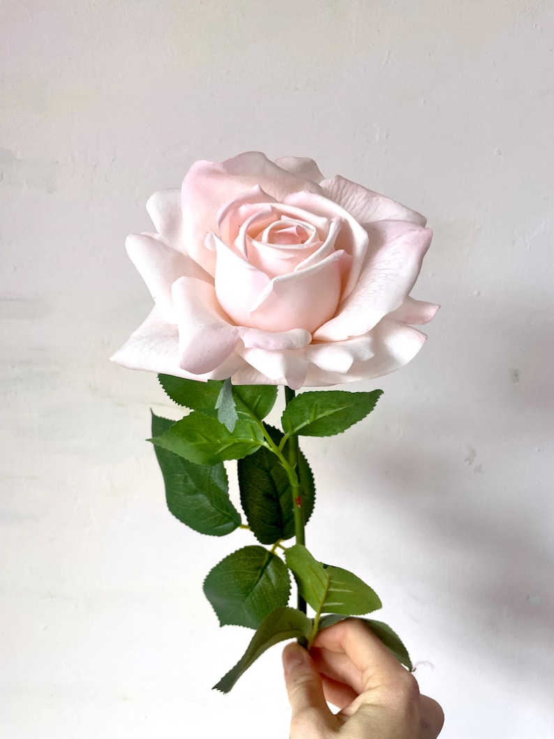 May include: A single, light pink artificial rose with green leaves and a stem.