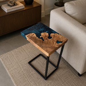 Puede incluir: Una mesa auxiliar rectangular con una superficie de madera natural de borde irregular y una incrustación de resina azul intenso. La mesa está sostenida por un marco de metal negro en forma de C, que proporciona una base estable a la madera.