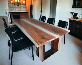 May include: A large wooden dining table with a white epoxy resin inlay. The table has a modern design with a rectangular shape and a unique base. The table is surrounded by black chairs.
