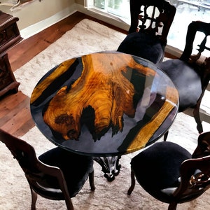 May include: A round wooden table with a black epoxy resin top. The table has a natural wood grain pattern and is surrounded by four black chairs with ornate backs.
