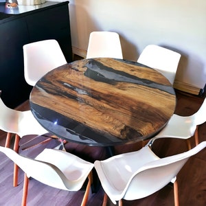 May include: A round wooden table with a black epoxy resin inlay. The table is surrounded by six white chairs.