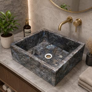 May include: A square vessel sink crafted from dark blue and gray stone with a natural, textured, crystalline appearance. The basin features a central circular drain with a silver-toned metal rim, set against a neutral stone countertop.