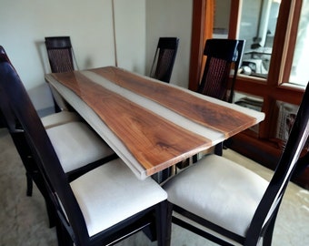 May include: A dining table with a live edge wooden top and a white epoxy resin inlay. The table is surrounded by six black chairs with white cushions.