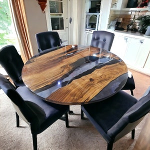 May include: A round wooden dining table with a black epoxy resin inlay. The table is surrounded by six black chairs with upholstered seats.