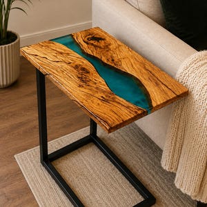 May include: A C-shaped side table featuring a rectangular top made from two pieces of natural wood with a prominent grain and knots, joined by a central river of translucent teal blue resin, supported by a black metal frame base.