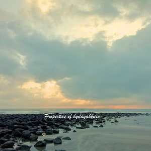 May include: A scenic coastal landscape featuring a beach with dark, rounded rocks and a calm sea under a cloudy sky. The text "Properties of bydayahburn" is visible. The sky transitions from a pale yellow to a muted blue, creating a serene atmosphere.