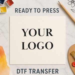 Könnte beinhalten: Ein quadratischer DTF-Transferbogen mit dem schwarzen Text YOUR LOGO zentriert auf einem cremefarbenen Hintergrund. Der Text READY TO PRESS steht über dem Quadrat und DTF TRANSFER darunter, umgeben von Aquarellfarben und einem Pinsel.