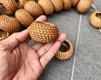以下が含まれることがあります： 自然色のラタンで作られた小さな織りバスケットが手に持たれています。バスケットは丸みを帯びた形で、織り模様があります。背景には他の同様のバスケットがいくつかあり、装飾品のセットまたはコレクションを示唆しています。