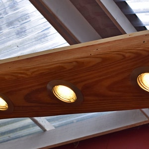 Könnte beinhalten: Eine rechteckige Holzleuchte mit vier Einbaustrahlern. Die Leuchte wird von dünnen Drähten von der Decke abgehängt. Das Holz hat eine natürliche Maserung und eine warme braune Farbe, wobei die Lichter ein sanftes Leuchten ausstrahlen.