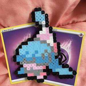 Puede incluir: Un personaje de pixel art azul, rosa, blanco y gris hecho de pequeñas cuentas de plástico. El personaje está sentado en una carta de intercambio de Pokémon con la palabra "ENERGY" impresa en ella.