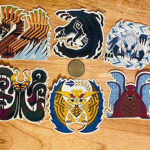 Può includere: Una collezione di sei adesivi colorati con disegni di mostri stilizzati. Gli adesivi presentano varie creature in tonalità di blu, rosso, giallo e marrone, con dettagli intricati e contorni audaci. Gli adesivi sono su una superficie di legno.