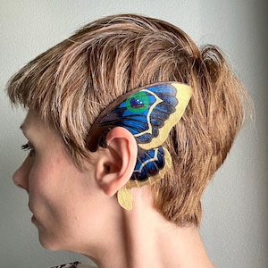 May include: A decorative ear cuff featuring a butterfly wing design. The wing is painted with blue, gold, and green hues, and is attached to a brown base that curves around the ear. The cuff is designed to be worn on the ear.