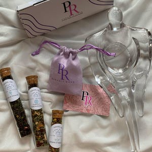 Peut inclure: Trois flacons en verre avec bouchons en liège remplis d'herbes séchées et étiquetés avec le texte "Paradis Rose" et des descriptions des bienfaits des herbes. Les flacons sont sur une surface blanche avec un sac à cordon violet, une carte rose avec le texte "PR Paradis Rose" et une sculpture en verre transparent.