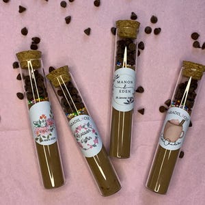 Könnte beinhalten: Vier Reagenzgläser aus Glas, gefüllt mit Schokoladenstückchen und Streuseln. Jedes Reagenzglas hat ein Etikett mit einem anderen Namen und Datum. Die Etiketten sind mit Blumen und Herzen verziert. Die Reagenzgläser befinden sich auf einem rosa Hintergrund.