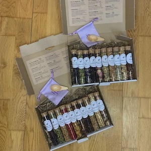 Peut inclure: Plusieurs boîtes en carton ouvertes remplies de tubes en verre contenant diverses herbes et épices séchées. Chaque tube est muni d'un bouchon en liège et d'une étiquette. De petits sachets violets avec des cuillères en bois sont également inclus.