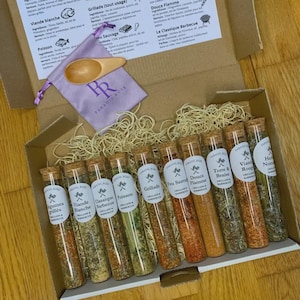Op de afbeelding: Een cadeaupakket met kruidenmengsels in glazen buisjes met kurken, gerangschikt in een kartonnen doos. De set bevat een kleine houten lepel, een paars zakje en etiketten met tekst. De buisjes bevatten verschillende kleuren kruiden.