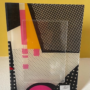 Peut inclure: Un cadre photo rectangulaire avec un design géométrique coloré. Le cadre présente un motif à pois noirs et blancs, avec des formes géométriques roses, jaunes et noires. Un insert en plastique transparent est au centre. Le cadre porte la signature "Albini".