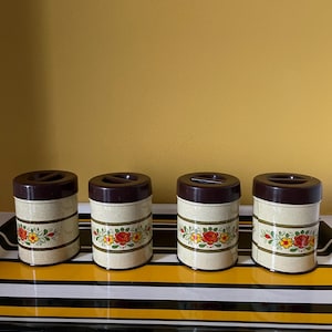 Peut inclure: Quatre bocaux vintage avec des couvercles marron et un corps beige. Chaque bocal présente un motif floral avec des roses rouges et jaunes. Les bocaux sont disposés sur un plateau à rayures noires, blanches et jaunes.