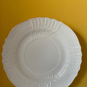May include: A white, round ceramic plate with a decorative, scalloped edge. The plate features a textured border and wave-like embossed designs. The plate is set against a yellow background.